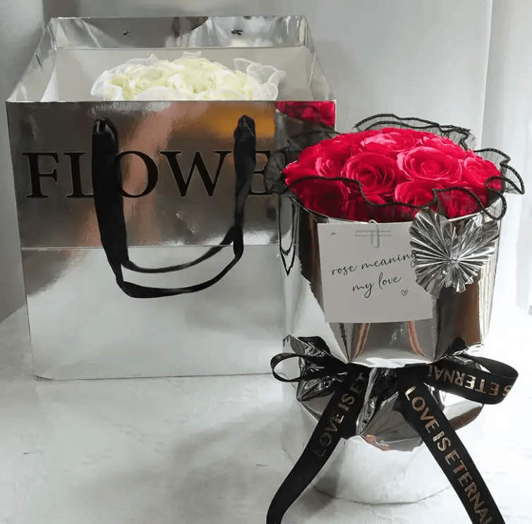 Elegant soap flower bouquet gift in a stylish metallic bag, featuring red roses and white flowers with a decorative tag.