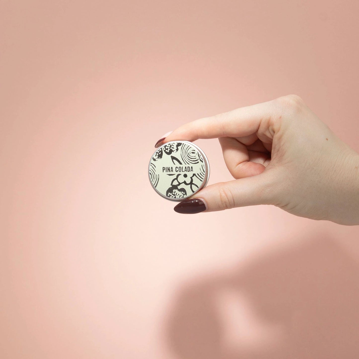Hand holding Agnes + Cat Pinacolada lip balm container against a soft pink background.
