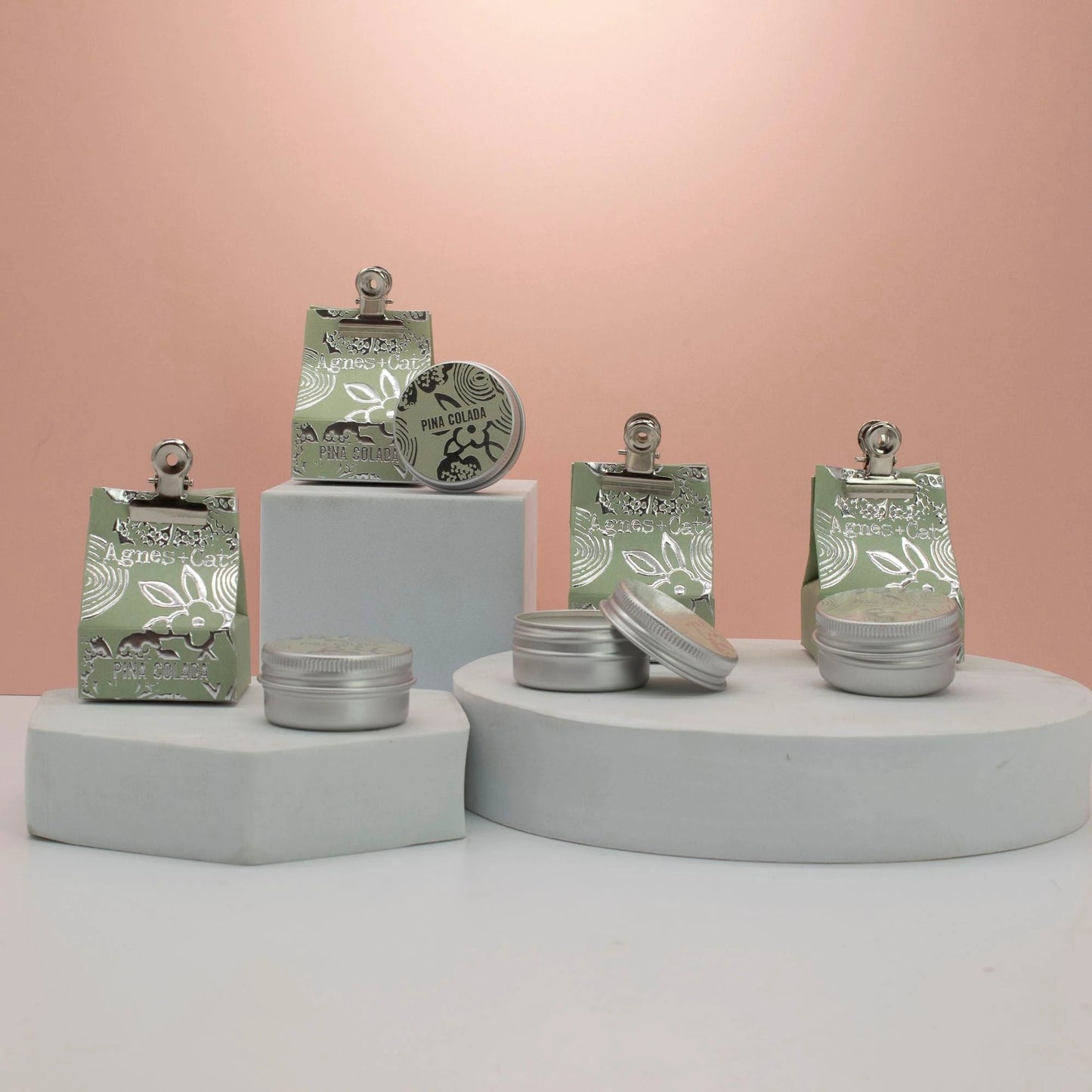 Agnes + Cat Pinacolada lip balm displayed in stylish containers on geometric pedestals with a pastel background.