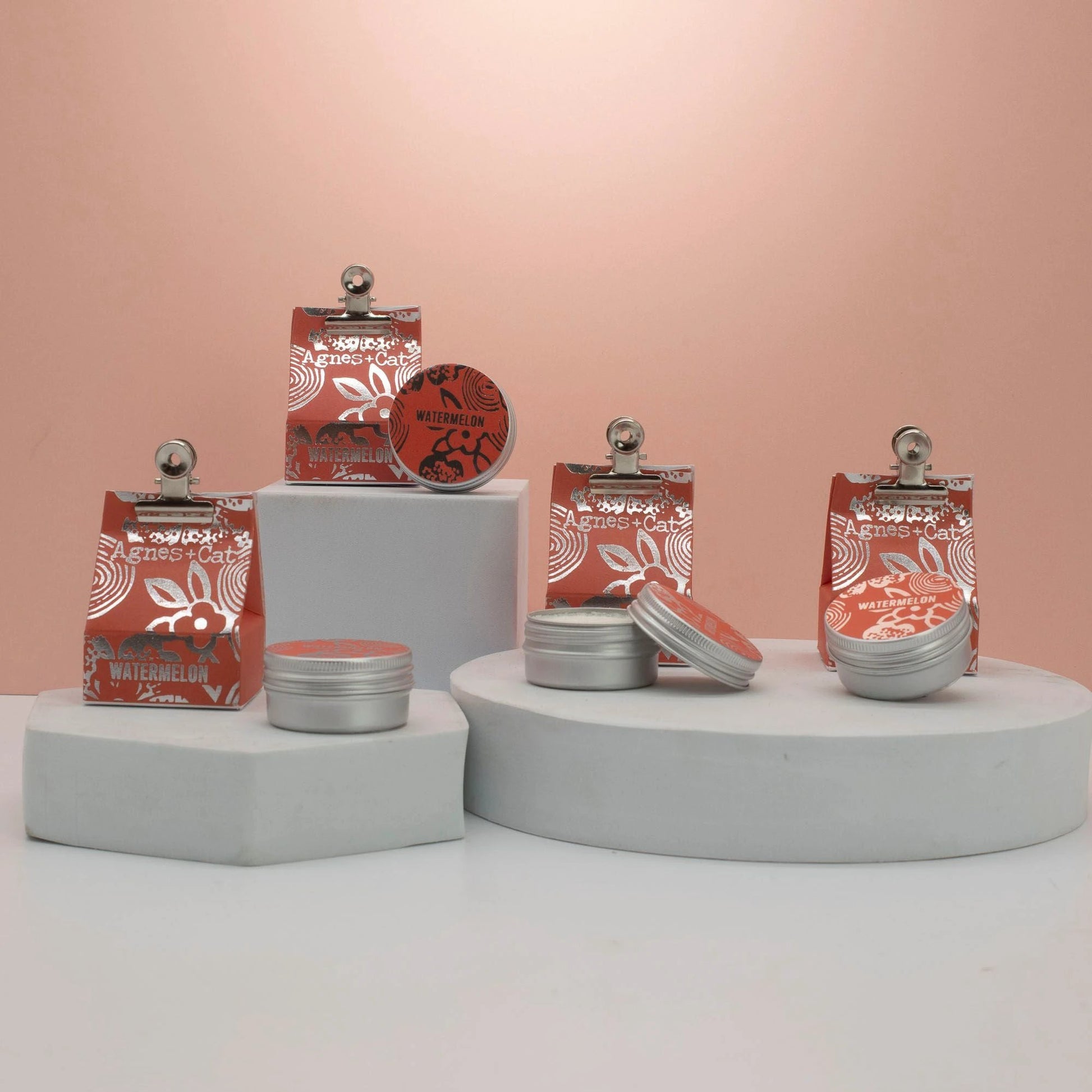 Agnes + Cat watermelon lip balm UK displayed in compact aluminium tins on pedestals against a soft pink background.